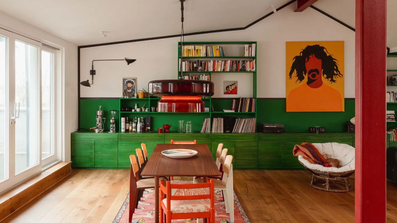 Colorful open-plan dining room, green painted cabinetry and shelving, dark wooden dining table with colorful dining chairs, warm wood floor