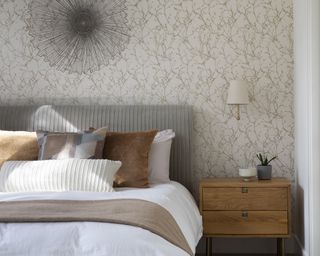 Bedroom with bed dressed with white linen and neutral throw and pillows, nightstand, wall light, patterned wallpaper and wall decor